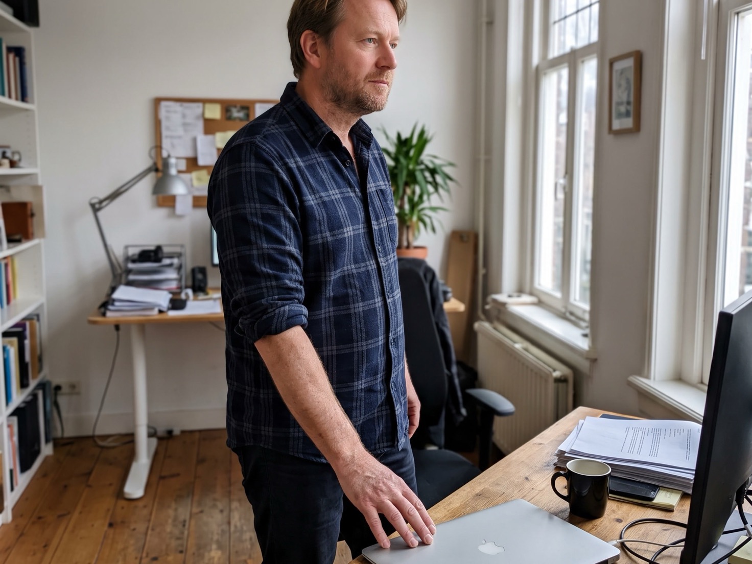 Man van rond de vijftig staat naast zijn bureau in een licht thuiskantoor, met de laptop dichtgeklapt