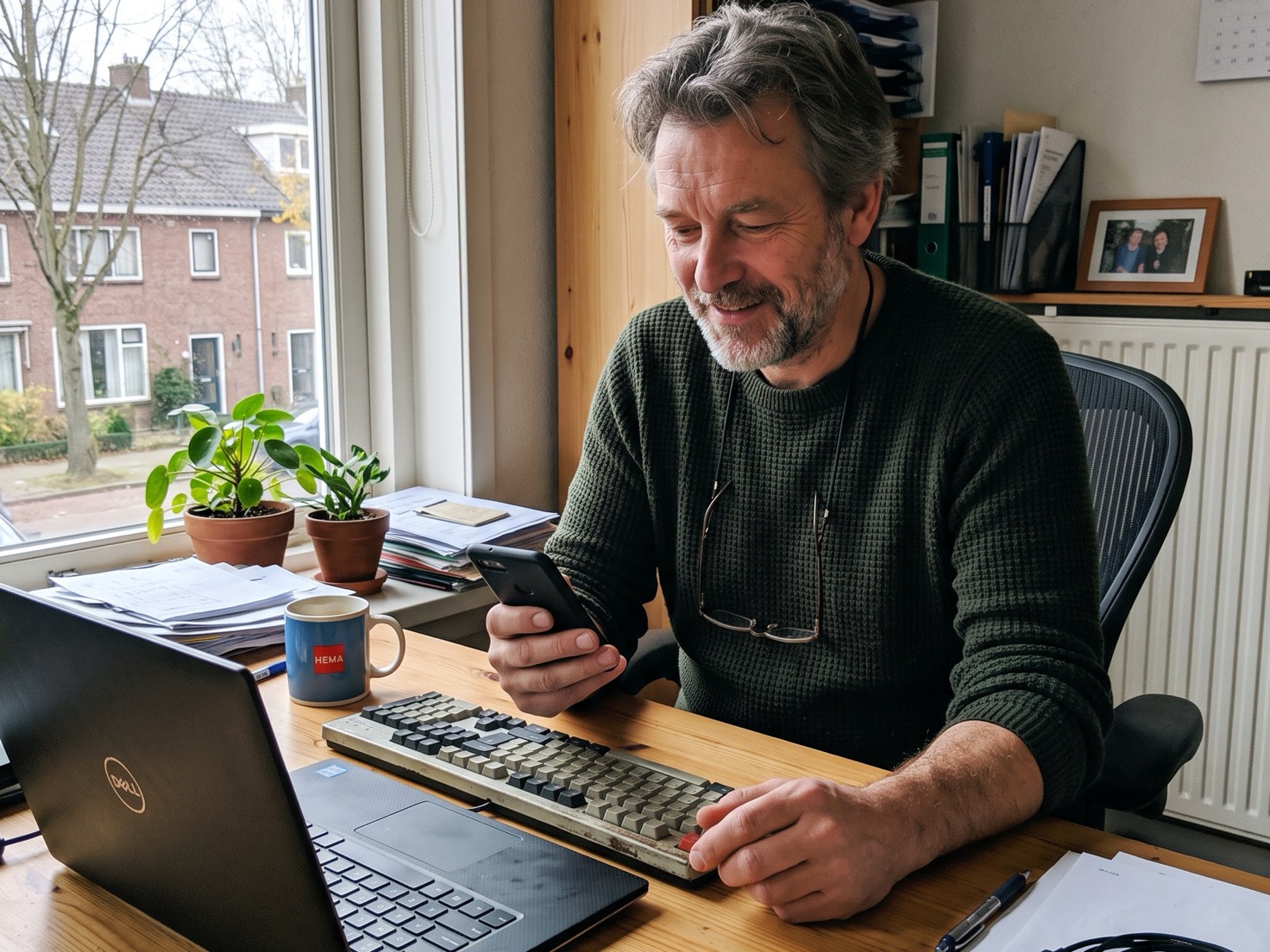 Man van middelbare leeftijd kijkt op zijn telefoon naast een open laptop aan zijn bureau thuis