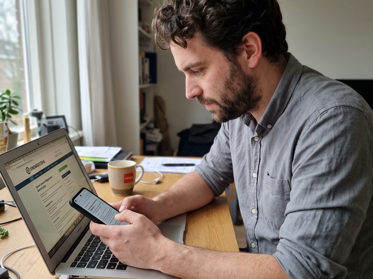 Zelfstandig ondernemer checkt op zijn telefoon de domeinregistratie van zijn website, laptop ernaast open op zijn eigen site