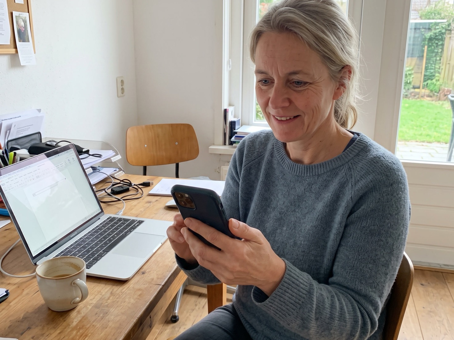 Vrouw kijkt glimlachend op haar telefoon aan haar bureau, met laptop en koffie ernaast