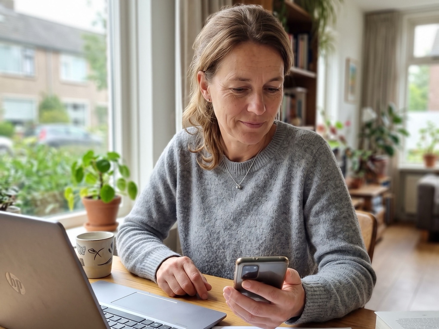 Vrouw van begin vijftig aan haar werktafel, laptop open naast zich, telefoon in de hand