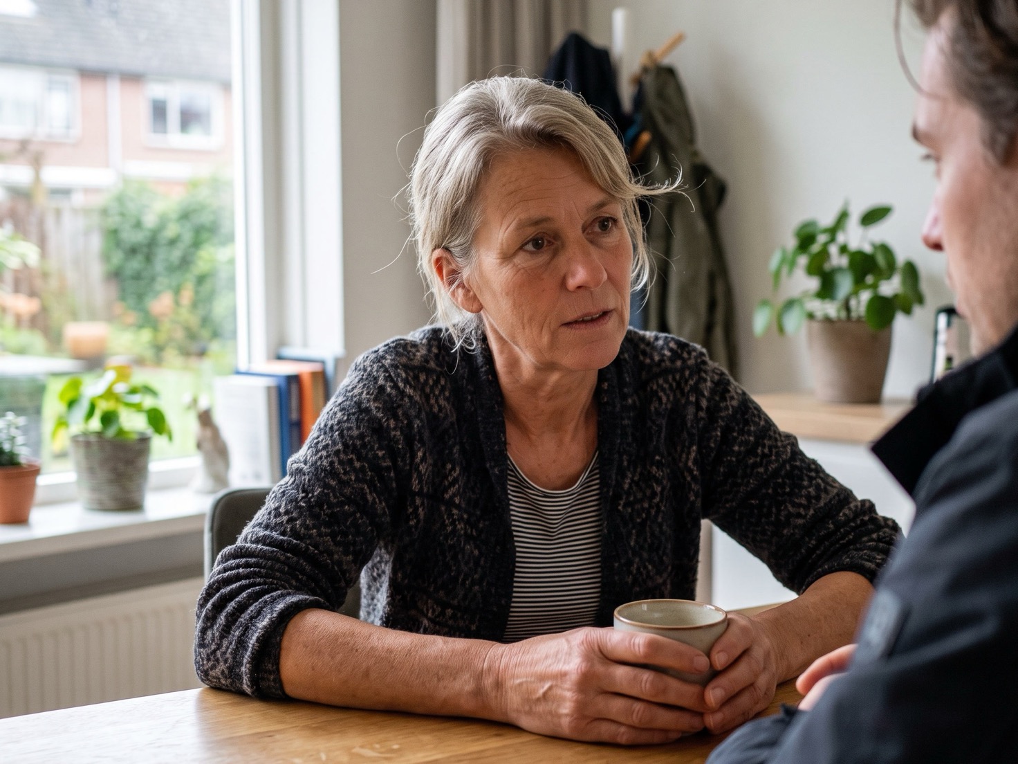 Vrouw met een kop koffie aan de eettafel in gesprek met iemand tegenover haar