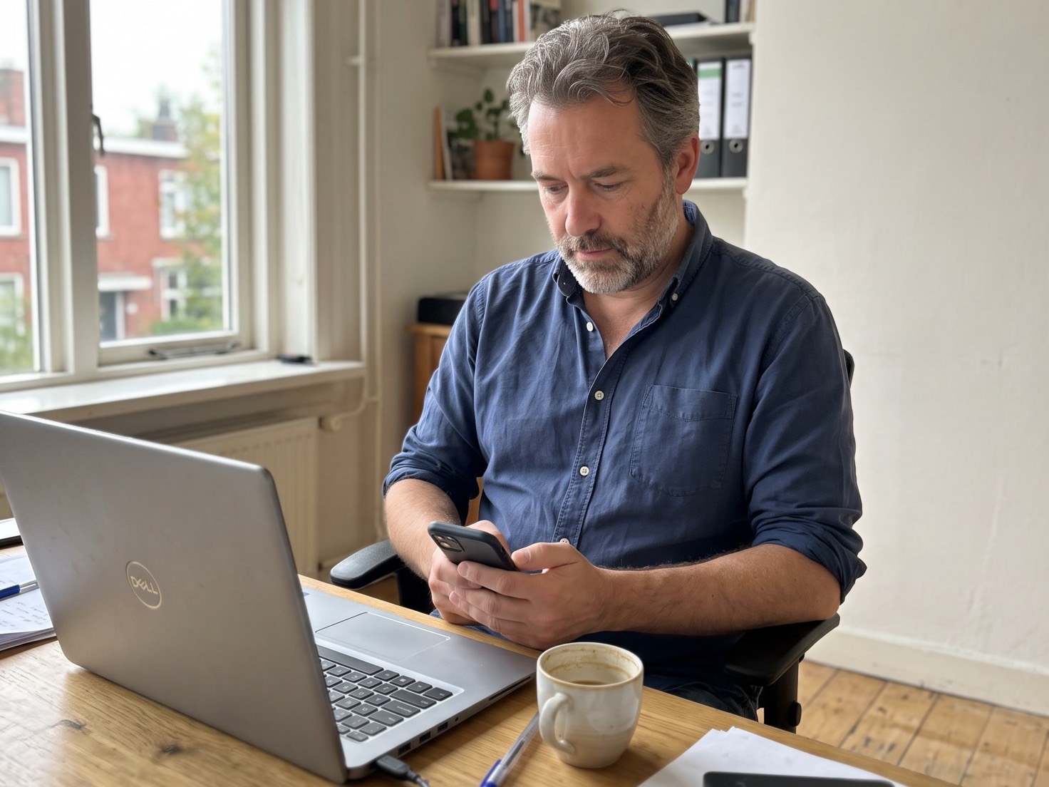 Zelfstandig ondernemer checkt zijn telefoon naast een openstaande laptop op zijn werkplek