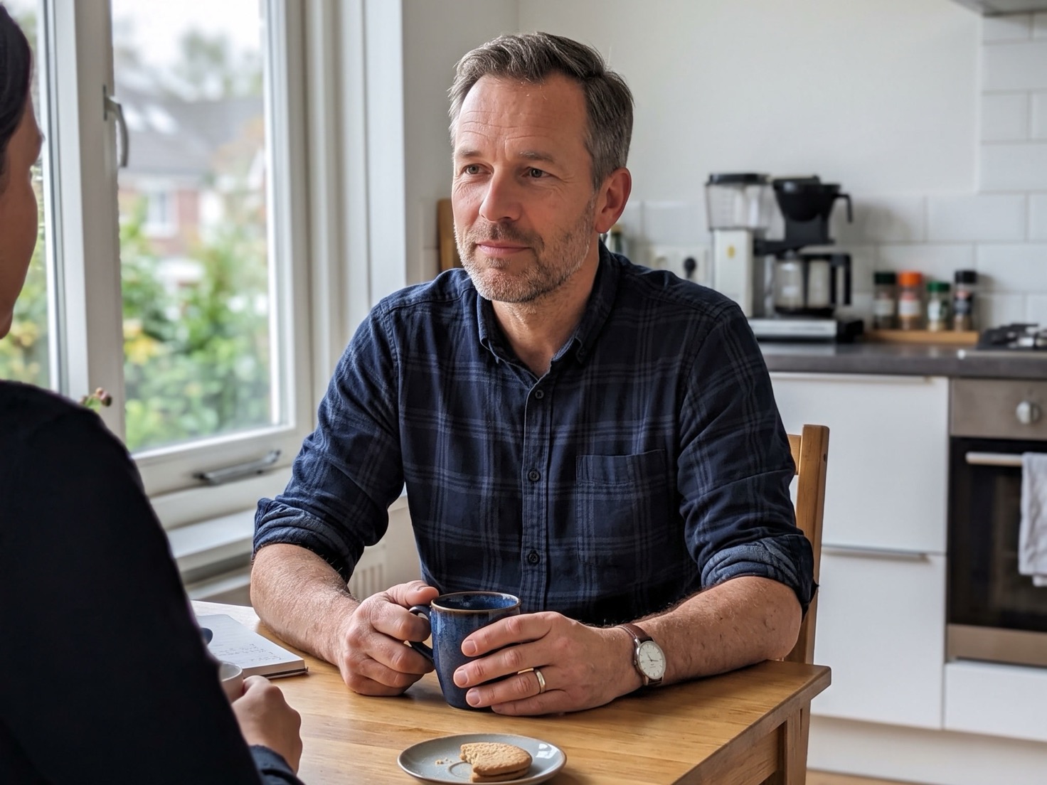 Ondernemer aan de keukentafel in gesprek met een klant, met een koffiemok in beide handen