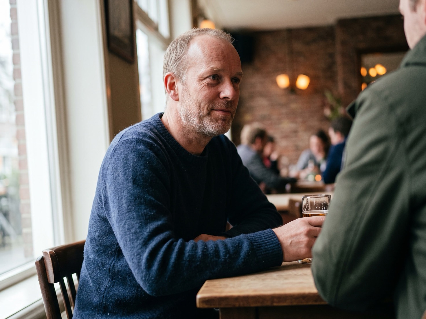 Man van rond de vijftig aan tafel in gesprek met iemand tegenover hem, bij daglicht thuis